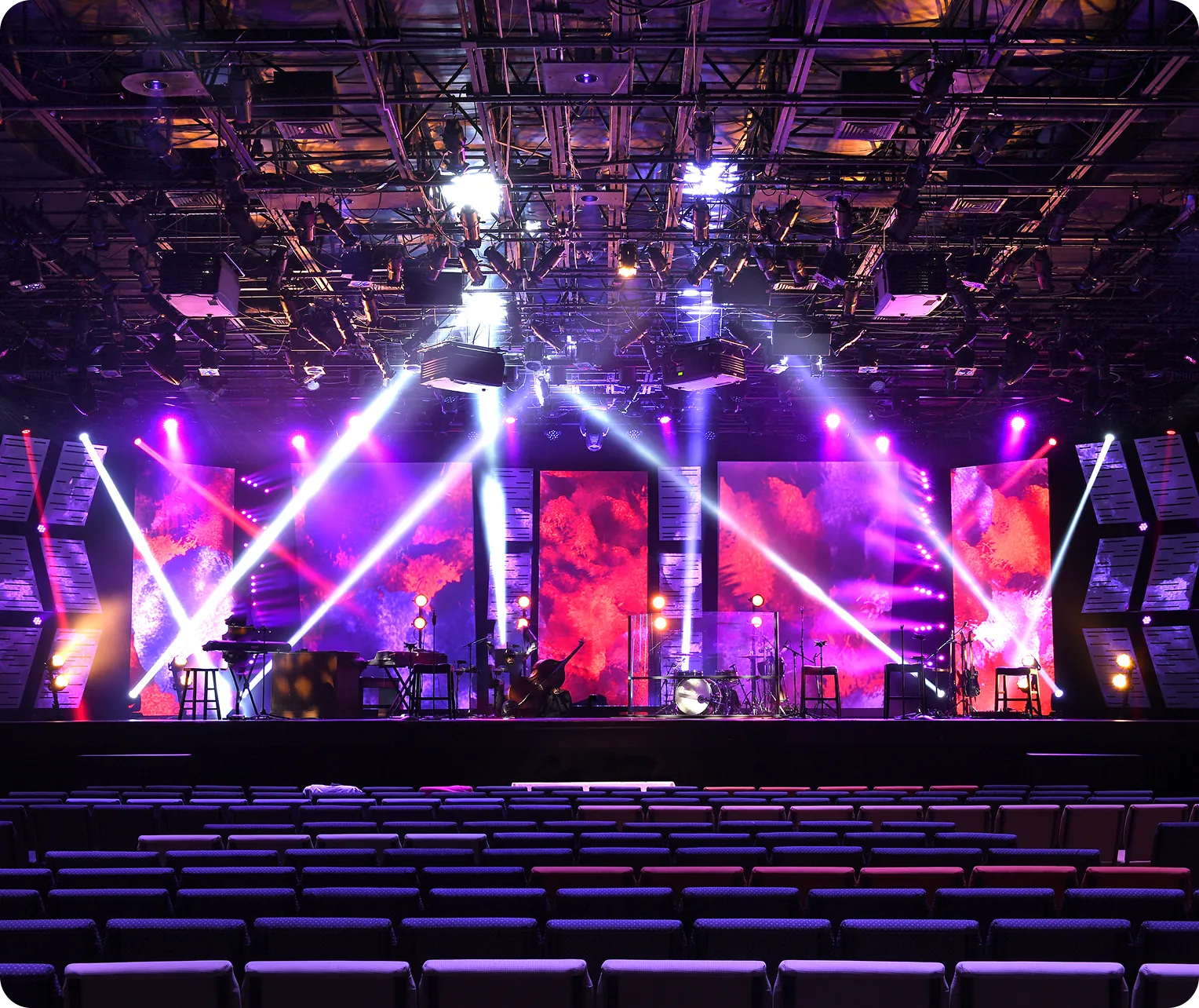 Concert stage with vibrant purple lighting, multiple spotlights, and various musical instruments set up. Background features colorful, abstract light displays.