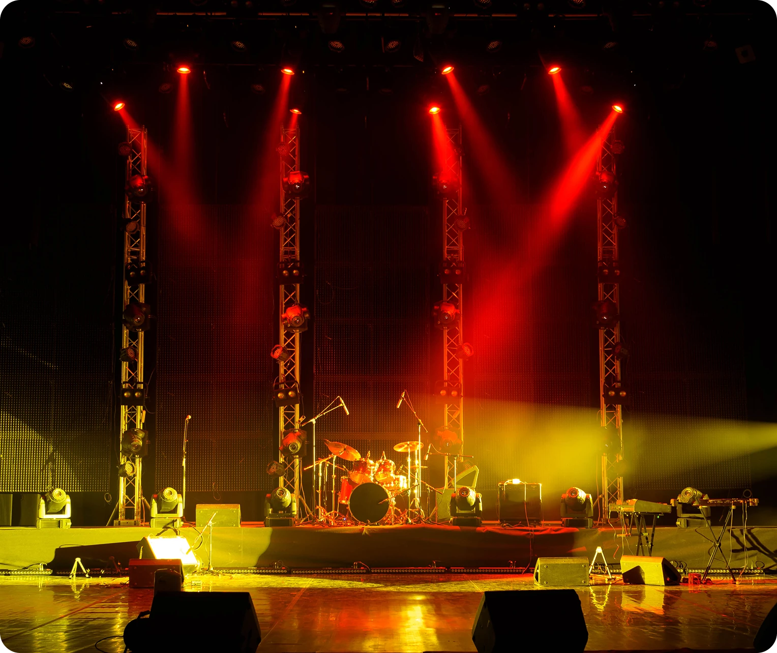 A dimly lit stage with red lights features a drum set and several microphones, with amplifiers and music equipment in the background.
