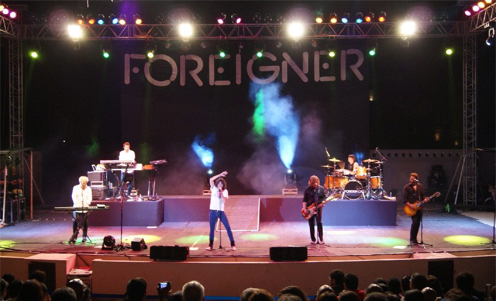 A band performing on stage with instruments and colorful lights, the backdrop displays the word "FOREIGNER.